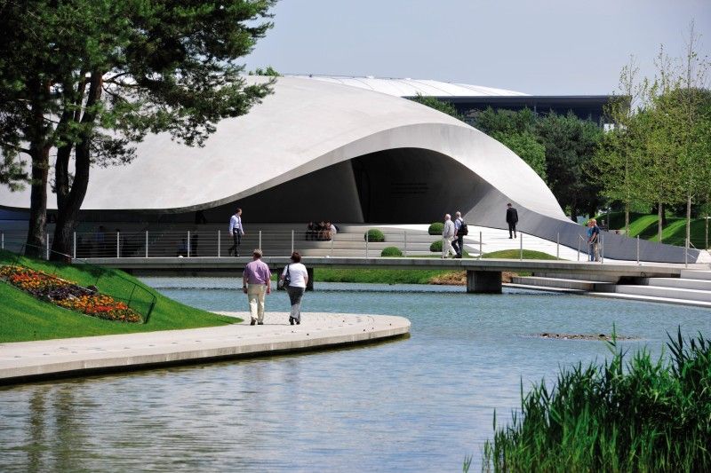 Zeitgenössisches Gebäude in natürlicher Umgebung Moderne Architektur mit geschwungenen Formen, umgeben von einem Teich und Grünanlagen. Menschen spazieren auf Wegen und über eine Brücke.